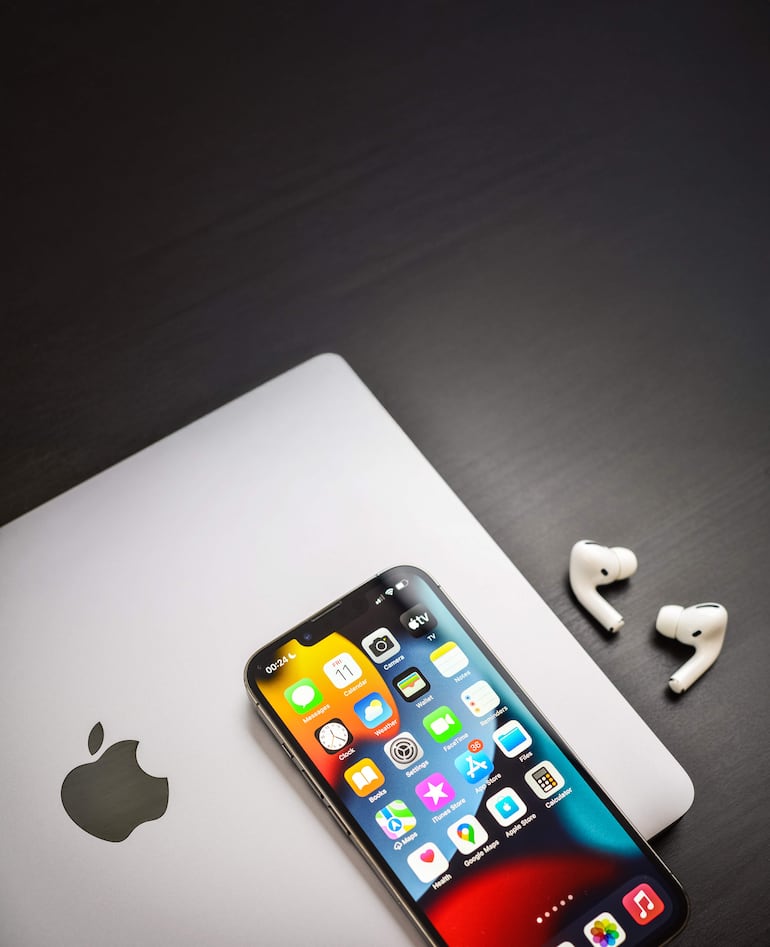 Laptop Apple con logotipo visible, teléfono móvil y auriculares inalámbricos sobre superficie oscura en un ambiente interior.