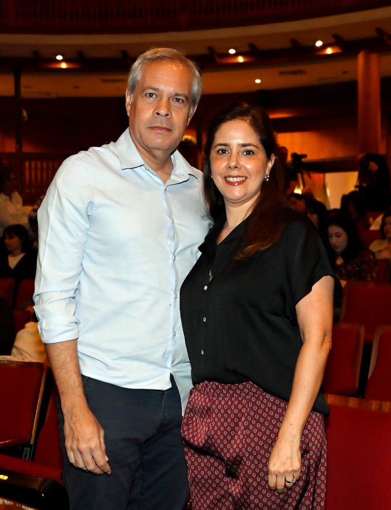 Hombre con camisa clara y pantalones oscuros junto a mujer en blusa negra y pantalones estampados, sonriendo en un teatro.