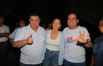 Tres personas sonrientes en ambiente nocturno, una levanta el pulgar, otra muestra paz con dos dedos y otra lleva camiseta con 'ES SOL'.