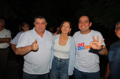 Tres personas sonrientes en ambiente nocturno, una levanta el pulgar, otra muestra paz con dos dedos y otra lleva camiseta con 'ES SOL'.