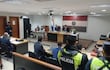 Tribunal con tres jueces al frente, hombre con uniforme policial de seguridad, personas atentas en ambiente de juicio.