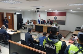 Tribunal con tres jueces al frente, hombre con uniforme policial de seguridad, personas atentas en ambiente de juicio.