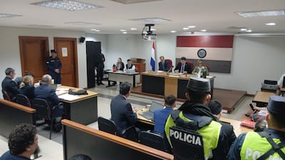Tribunal con tres jueces al frente, hombre con uniforme policial de seguridad, personas atentas en ambiente de juicio.