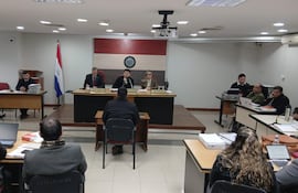 Tres miembros del panel en sala de audiencias, observando atentamente mientras un hombre, de espaldas, expresa su testimonio.