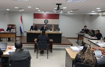 Tres miembros del panel en sala de audiencias, observando atentamente mientras un hombre, de espaldas, expresa su testimonio.