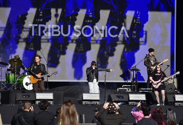 La cantante de Tribu Sónica, con micrófono en mano, se destaca en el escenario mientras el guitarrista toca al centro.
