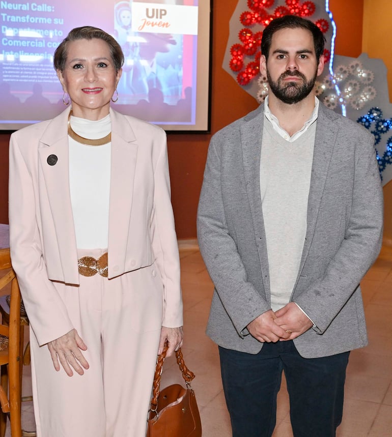 Mujer con blazer claro y bolso marrón, junto a hombre con chaqueta gris, en un evento decorado y formal.