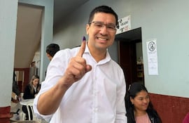 Hombre con camiseta blanca y gafas sonríe mientras levanta el dedo índice marcado con tinta en un lugar de votación.