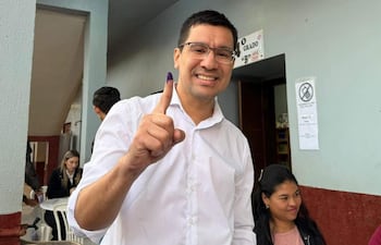 Hombre con camiseta blanca y gafas sonríe mientras levanta el dedo índice marcado con tinta en un lugar de votación.
