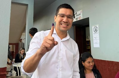 Hombre con camiseta blanca y gafas sonríe mientras levanta el dedo índice marcado con tinta en un lugar de votación.