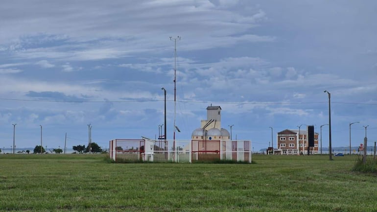 Campo verde con cancha de fútbol y arco blanco, edificios en el fondo bajo un cielo gris.