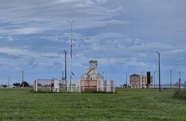 Campo verde con cancha de fútbol y arco blanco, edificios en el fondo bajo un cielo gris.