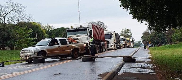 Policía en uniforme junto a camioneta pequeña, controlando camiones detenidos en carretera mojada rodeada de vegetación.
