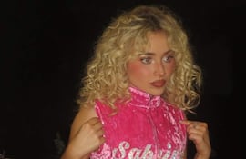 Sabrina Carpenter con vestido rosa con el nombre 'Sabrina' en brillante, en ambiente oscuro, luciendo seria y concentrada.