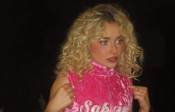 Sabrina Carpenter con vestido rosa con el nombre 'Sabrina' en brillante, en ambiente oscuro, luciendo seria y concentrada.