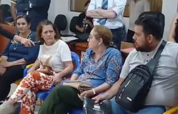 Mujeres sentadas en sillas amarillas, atendiendo en un ambiente cerrado. Hombre a la derecha sostiene una cartera negra.
