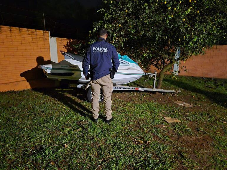 Oficial de policía de espaldas en jardín con chaqueta 'POLICÍA INVESTIGACIONES', junto a moto de agua Yamaha.