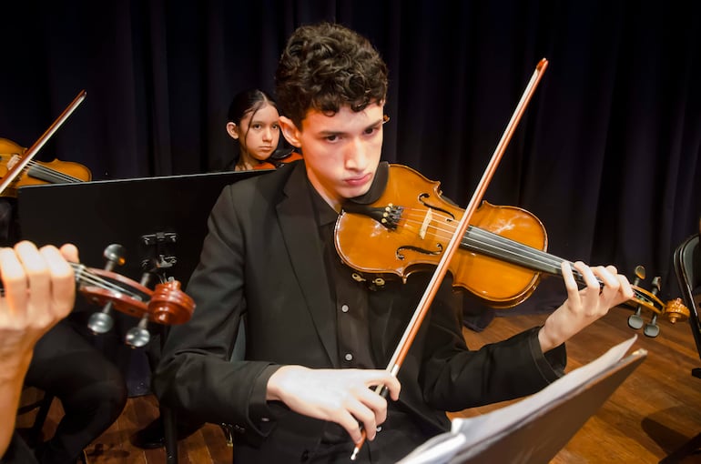 Joven músico con cabello rizado sostiene un violín, mientras otra joven lo escucha atentamente en un escenario oscuro.