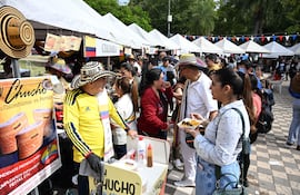 Hombre con sombrero colombiano y camiseta amarilla vende comida, mientras una mujer interactúa con él en un ambiente festivo.