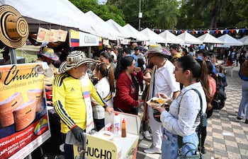Hombre con sombrero colombiano y camiseta amarilla vende comida, mientras una mujer interactúa con él en un ambiente festivo.