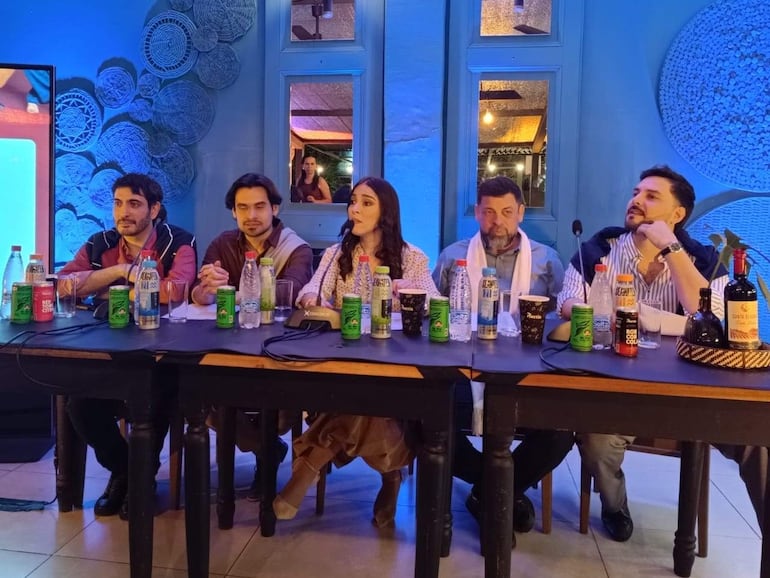 Cinco personas sentadas en mesa: mujer de vestimenta clara habla, hombres escuchan atentos, ambiente moderno con botellas sobre la mesa.