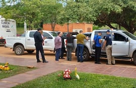 Grupo de seis personas en entorno suburbanos, entre ellas un policía y una mujer con blusa roja, junto a dos vehículos.