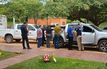 Grupo de seis personas en entorno suburbanos, entre ellas un policía y una mujer con blusa roja, junto a dos vehículos.