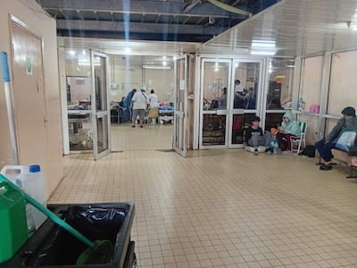 Pacientes sentados en el suelo y sillas, con profesionales de salud atendiendo en el fondo, en un hospital con luz artificial.