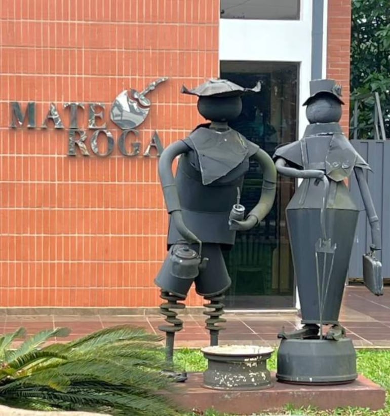 Esculturas de metal de pareja sosteniendo objetos relacionados con el mate, en un entorno natural y acogedor.
