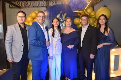 Tres hombres en trajes oscuros y tres mujeres en vestidos de fiesta, sonriendo en un fondo decorado en dorado y azul.