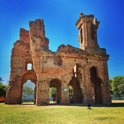 Estructura en ruinas con ladrillos rojizos y arcos góticos, rodeada de césped verde en un día soleado.
