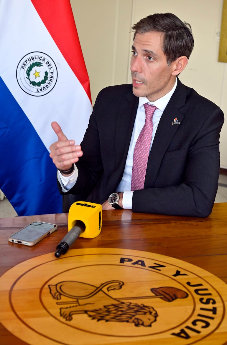 Marcos Riquelme en traje oscuro y corbata rosada gesticula durante la reunión con una bandera de Paraguay de fondo.