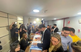 Mujer detrás de escritorio con gafas atiende a un hombre en traje oscuro, rodeados de periodistas en un ambiente interior.