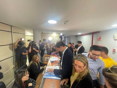Mujer detrás de escritorio con gafas atiende a un hombre en traje oscuro, rodeados de periodistas en un ambiente interior.