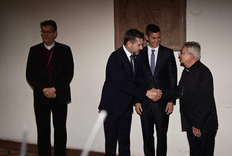 Cuatro hombres en trajes se dan la mano en un entorno formal; uno sonríe mientras observa, todos lucen tranquilamente atentos.