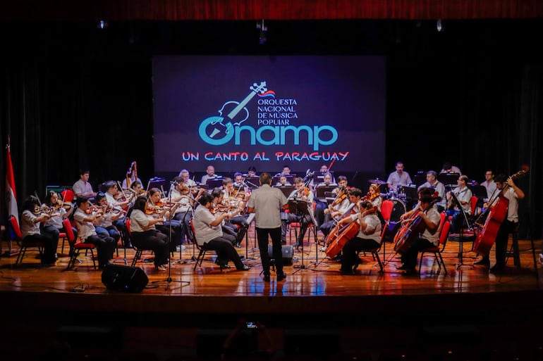 Músicos de la Orquesta Nacional de Música Popular en camisetas blancas, dirigidos por un hombre al frente en un auditorio con sillas rojas.