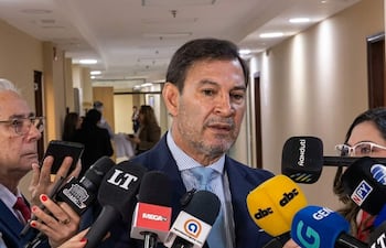 Silvio Ovelar rodeado de periodistas, con expresión seria y micrófonos de varios medios en un ambiente interno.