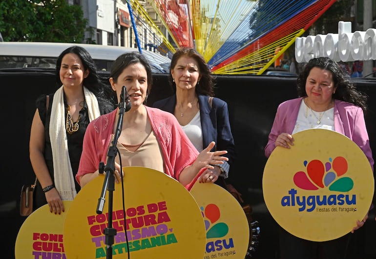 Mujer con micrófono y blusa clara ofrece discurso, mientras otras tres la observan con interés en evento colorido.