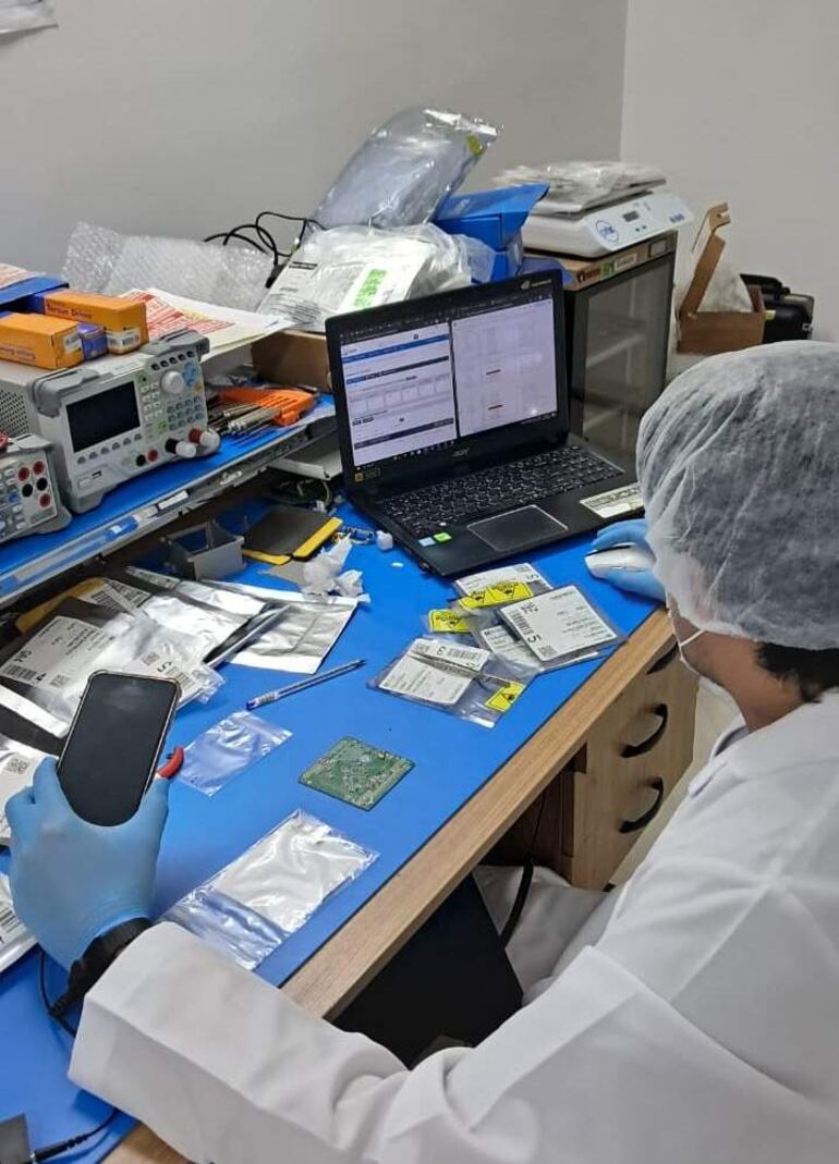 Técnico en bata blanca y guantes, enfocado en un teléfono, rodeado de herramientas y componentes electrónicos en un laboratorio desordenado.