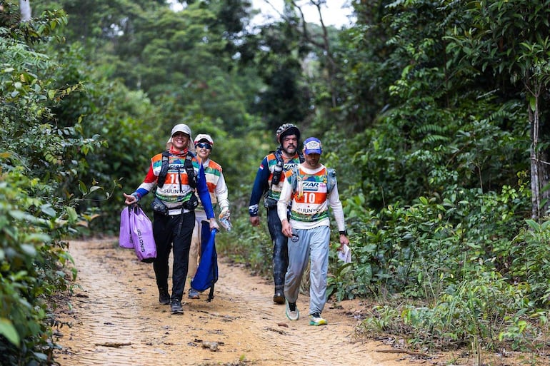 Competidores Angiru sonríen en el sendero, visten ropa deportiva y llevan mochilas en un entorno natural.