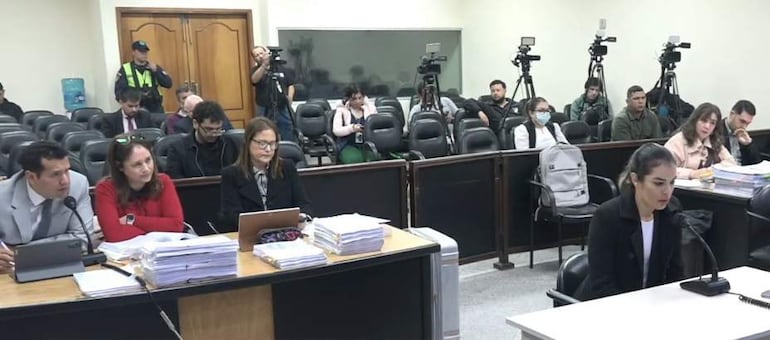 Tres mujeres sentadas alrededor de una mesa en una sala de audiencias, una de ellas con traje claro, mientras periodistas graban.