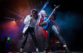 Mars Marshall con cabello rizado y vestimenta blanca canta energéticamente, mientras Jennifer Batten en traje rojo toca guitarra azul.