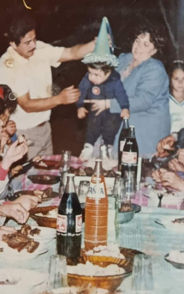 Niño con gorro de fiesta es sostenido por un adulto mientras otros miembros de la familia disfrutan en una mesa con comida y bebidas.