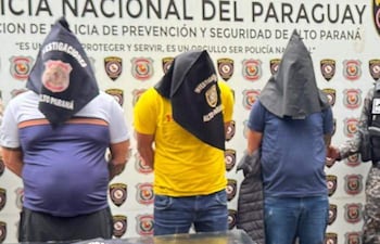 Tres hombres con togas oscuras de pie, frente al logo de la Policía Nacional del Paraguay, mientras un oficial supervisa la escena.
