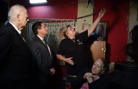 Hombres en trajes oscuros escuchan a mujer con cabello rizado, en ambiente doméstico con decoración roja.