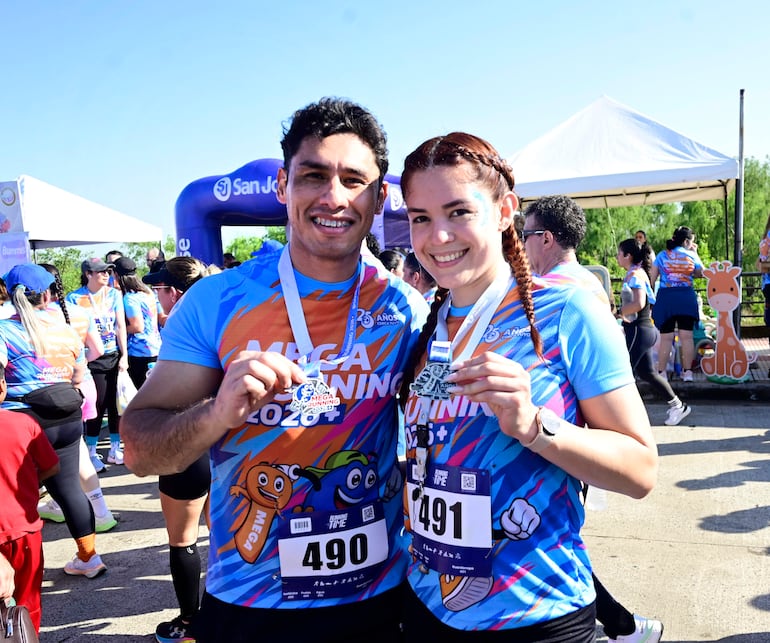 Hombre y mujer sonrientes, con camisetas coloridas y medallas, celebran en un evento de running al aire libre.