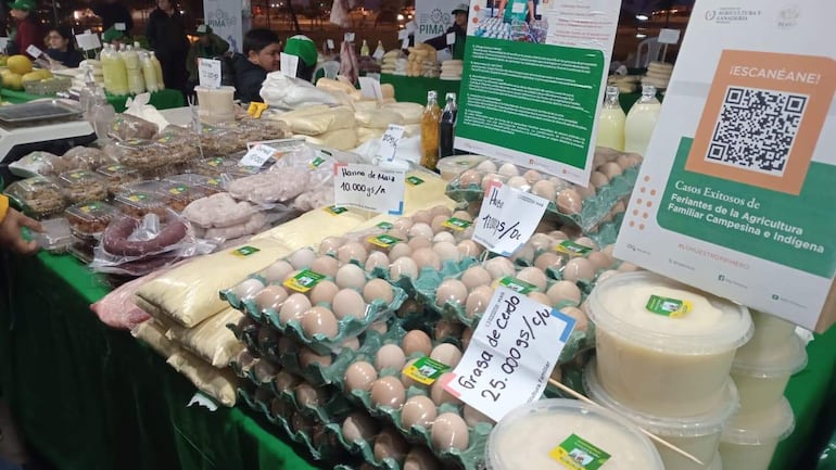 Mesa con productos agrícolas como huevos y harina de maíz exhibidos al aire libre en feria con luces de fondo.