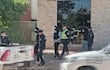 Tres hombres con pasamontañas son acompañados por dos policías en un ambiente urbano, frente a un edificio de ladrillos claros.