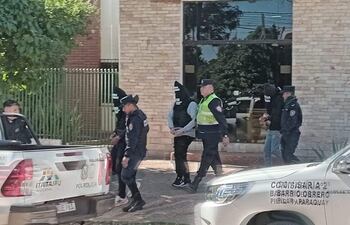 Tres hombres con pasamontañas son acompañados por dos policías en un ambiente urbano, frente a un edificio de ladrillos claros.