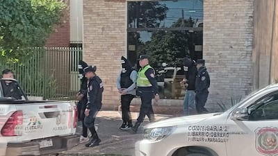 Tres hombres con pasamontañas son acompañados por dos policías en un ambiente urbano, frente a un edificio de ladrillos claros.